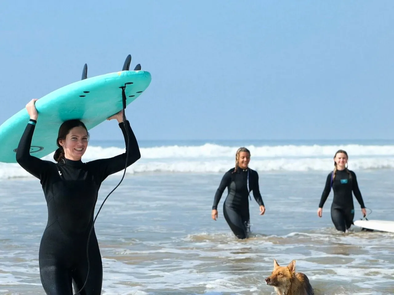 A surfer carrying her board on her head out of the sea, friends and a dog behind